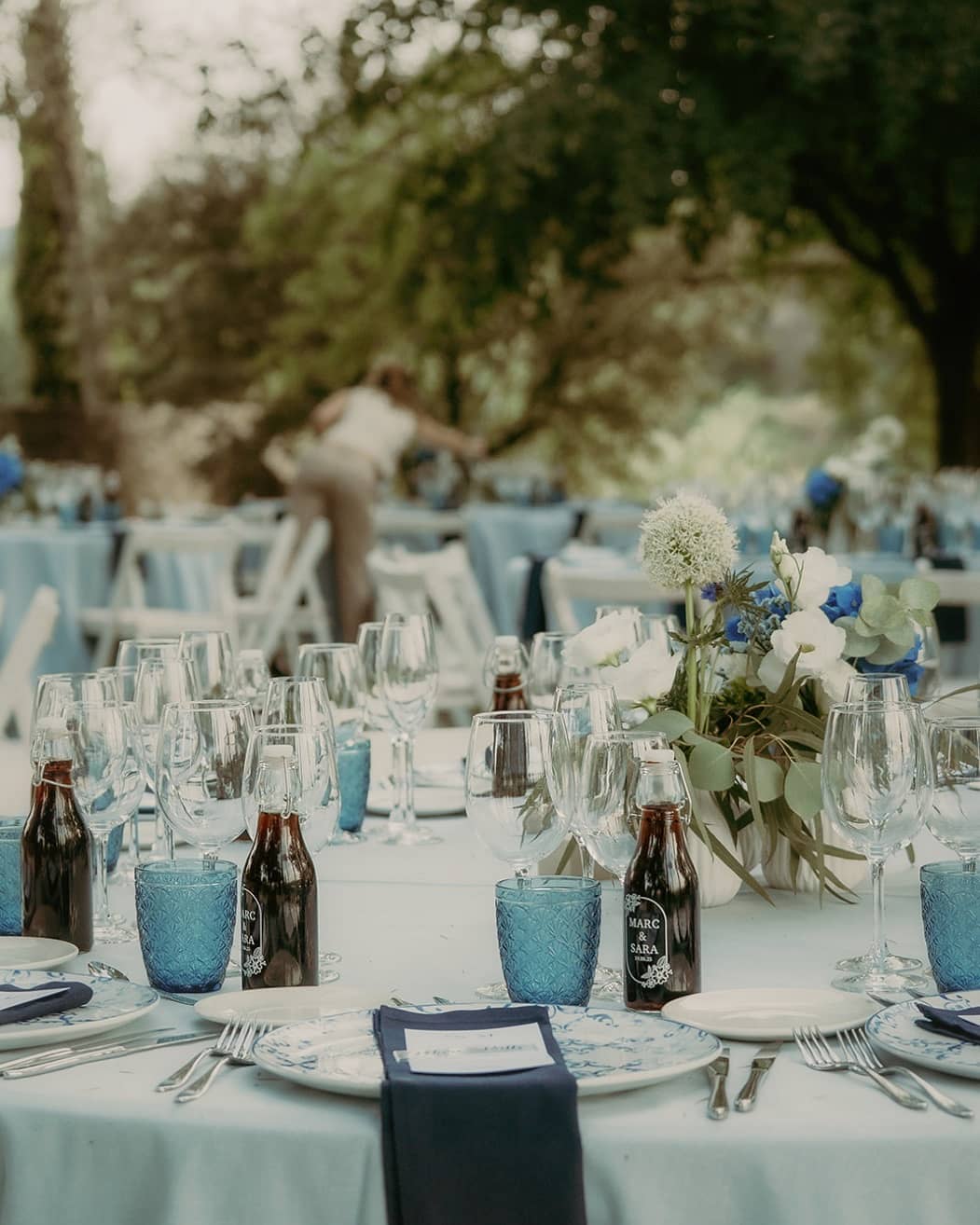 Mesa con mantel azul, vasos azules y centro de flores en un jardín
