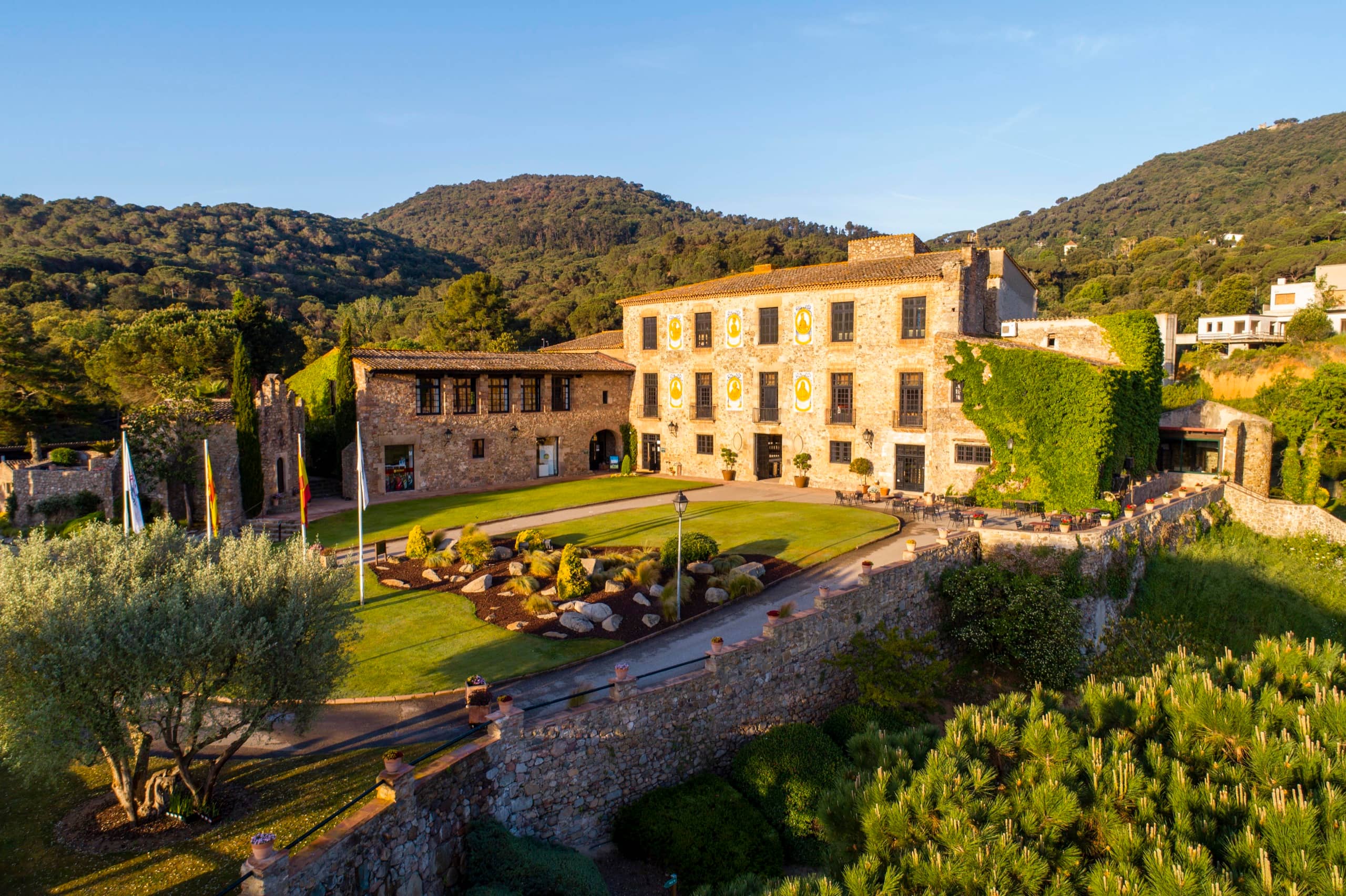 Masía de piedra rodeada de jardines y montañas al atardecer, vista desde el aire